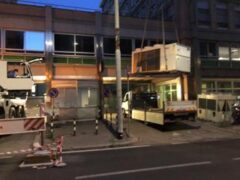 Camion con gru solleva container davanti a edificio.